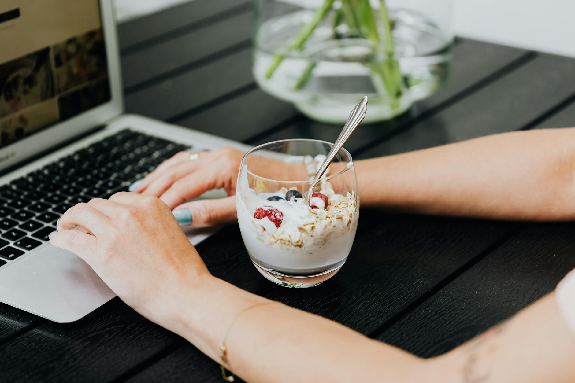 Osoba pracuje na laptopie, obok stoi szklanka z jogurtem i owocami