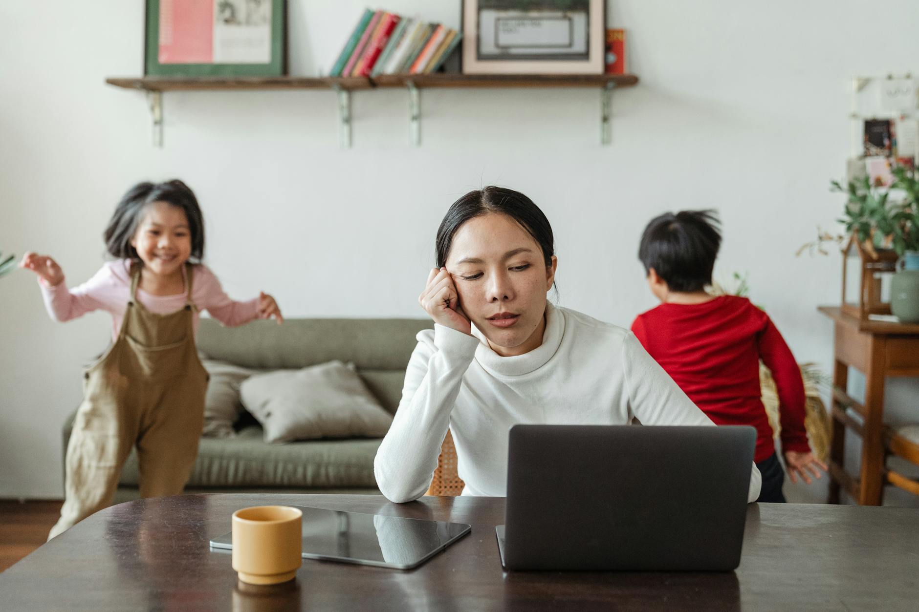 Zmęczona mama przy laptopie, dzieci bawią się obok w domu